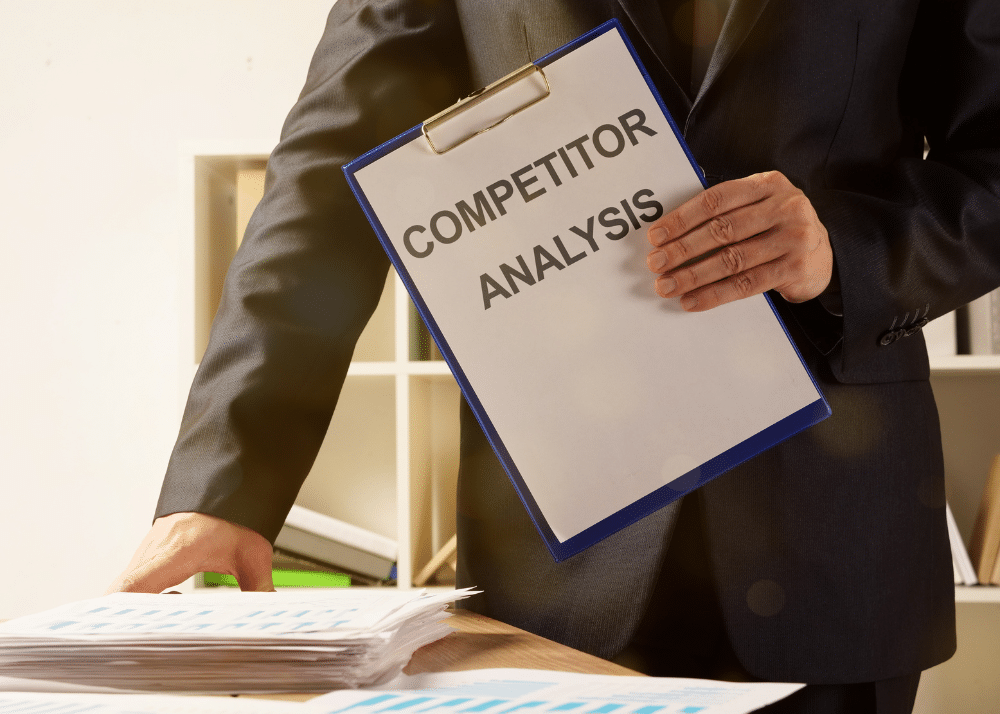 Eine Person in Business-Kleidung hält einen Clipboard mit der Aufschrift "COMPETITOR ANALYSIS" in der Hand, daneben liegt ein Stapel von Dokumenten.